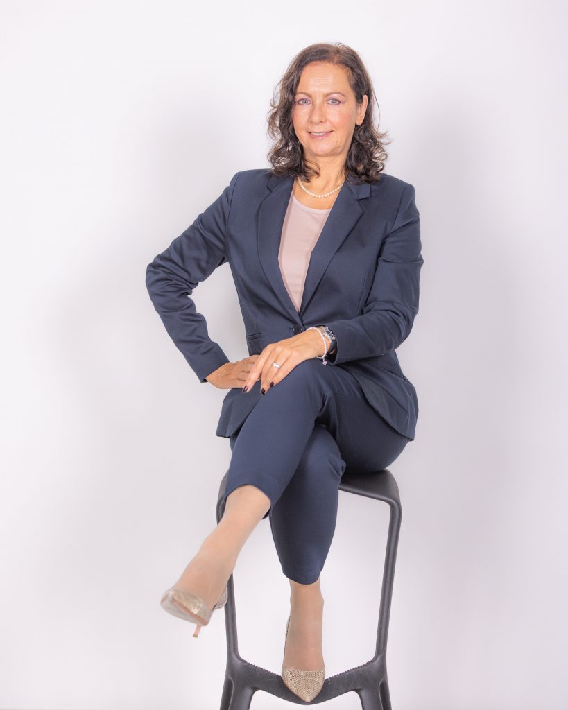 Karin Bacher sitzt in Business Kleidung auf einem Hocker